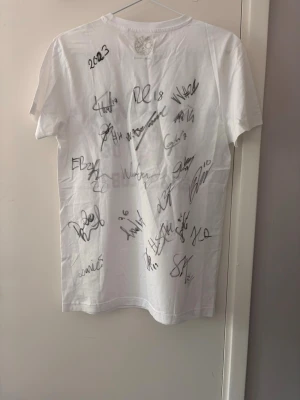 Örebro Sportklubb t-shirt med autografer - Vit t-shirt från Örebro Sportklubb med tryck framtill och flera autografer på baksidan. Klassisk rund hals och korta ärmar. Perfekt för dig som vill visa ditt stöd för laget!