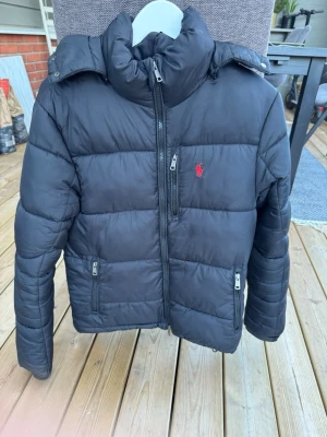 Svart dunjacka från Polo Ralph Lauren - Snygg svart dunjacka från Polo Ralph Lauren med hög krage och dragkedja framtill. Jackan har två framfickor med dragkedja och den klassiska röda Polo-loggan broderad på bröstet. Perfekt för kalla vinterdagar och riktigt stilren look.