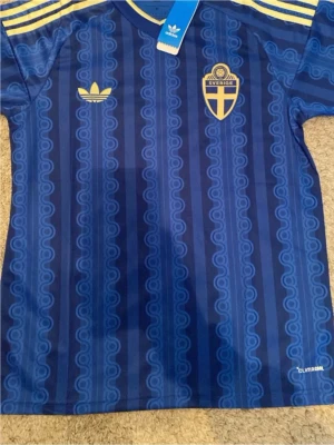 Blå Sverige fotbollströja Adidas - Säljer en blå fotbollströja från Adidas med Sveriges landslagsemblem på bröstet och gula detaljer. Tröjan har ett retroinspirerat mönster, gula ränder på axlarna och är tillverkad i lätt och ventilerande Climalite-material.