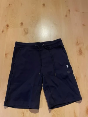 Mörkblå shorts från Polo Ralph Lauren - Snygga mörkblå shorts från Polo Ralph Lauren med klassisk logga broderad på ena benet. Shortsen har snörning i midjan och är tillverkade i mjuk bomull, perfekta för en avslappnad stil. Fickor på sidorna för extra funktionalitet.