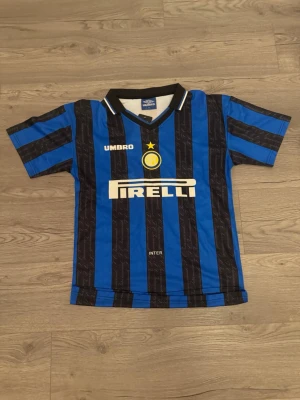 Inter Milan Ronaldo tröja Umbro - Klassisk Inter Milan fotbollströja från Umbro med blå och svarta ränder, Pirelli-logga på bröstet och Ronaldo 10 på ryggen. Tröjan har korta ärmar, krage med vit detalj och klubbmärke framtill. Perfekt för dig som älskar retro och fotboll.
