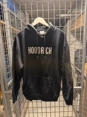 Svart HOODRICH hoodie med tryck - Svart HOODRICH sett med stor vit logga på bröstet och tryck på ena ärmen samt ryggen. Klassisk känguruficka framtill, dragsko i huvan och ribbade muddar. Perfekt för en streetwear-look.  Small i både tröja och Byxor 