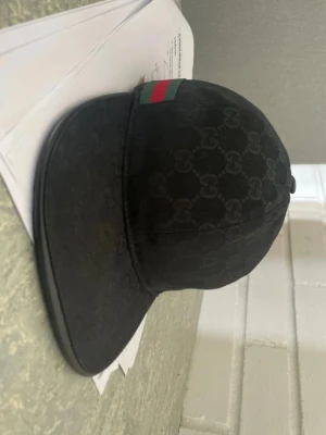 Svart keps från Gucci med GG-mönster - Snygg svart keps från Gucci med klassiskt GG-monogram över hela kepsen. Framtill finns en ikonisk grön och röd rand. Kepsen har böjd skärm och är tillverkad i tygmaterial. Perfekt för dig som vill ha en stilren och lyxig accessoar.