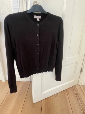 Svart kofta från H&M - Klassisk svart kofta från H&M med rund halsringning och knäppning framtill. Långärmad modell i mjukt material, perfekt att slänga över en t-shirt eller klänning. Enkel och stilren design som funkar till det mesta.