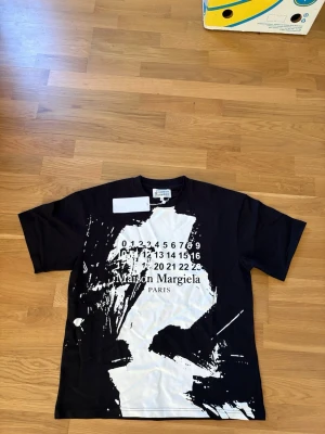 Maison Margiela svartvit t-shirt - Cool svart t-shirt från Maison Margiela med vit abstrakt print och ikonisk sifferlogga framtill. Oversized passform och klassisk rund hals. Perfekt statement-plagg för dig som gillar unika designer.