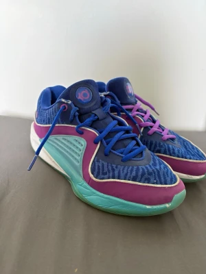 KD basketskor - Säljer ett par Nike KD basketdojor i blått, lila och turkost med vita detaljer. Skorna har mesh-överdel, lågt skaft och olika färg på skosnörena – ett par blå och ett par lila. Perfekta för dig som vill sticka ut på basketplanen. Skick 7,5/10. Kom privat för mer info!