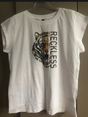 Vit t-shirt med tigertryck från PREPAIR - Snygg vit t-shirt från PREPAIR med ett coolt tigertryck och texten 'RECKLESS' på framsidan. T-shirten har rund halsringning och korta ärmar. Perfekt för dig som vill sticka ut med en trendig och bekväm look.