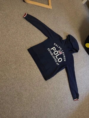 Marinblå hoodie från Polo Ralph Lauren - Snygg marinblå hoodie från Polo Ralph Lauren med stor vit logga och text framtill. Klassisk huva och långa ärmar med ribbade muddar, samt diskreta röda och vita detaljer vid ärmsluten. Perfekt för en avslappnad och stilren look.