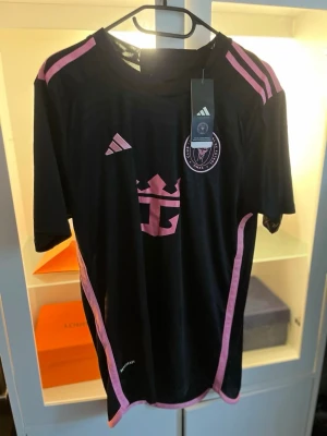 Svart Inter Miami matchtröja Adidas - Snygg svart matchtröja från Inter Miami med rosa detaljer och klubbmärke på bröstet. Tröjan har korta ärmar, rund hals och är tillverkad i ett lätt och ventilerande material. Perfekt för fotbollsträning eller att bära som supporter.