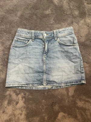 Blå jeanskjol i klassisk modell - Snygg blå jeanskjol i kort modell med klassiska fickor fram och bak samt bälteshällor. Kjolen har knapp och dragkedja framtill och är tillverkad i denim. Perfekt att matcha med en t-shirt eller hoodie.