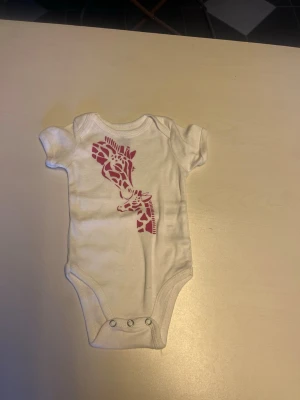 Vit body med girafftryck - Söt vit kortärmad body med ett rosa girafftryck på framsidan. Praktiska tryckknappar i grenen för enkel på- och avklädning. Perfekt för små barn och tillverkad i mjukt material som känns skönt mot huden.