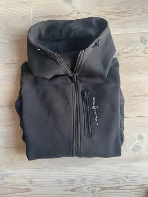 Svart hoodie från Sail Racing - Svart hoodie med dragkedja från Sail Racing. Hoodien har en bröstficka med dragkedja och logga tryckt på vänster sida. Klassisk passform med huva och ribbade muddar. Perfekt för en avslappnad och sportig stil.