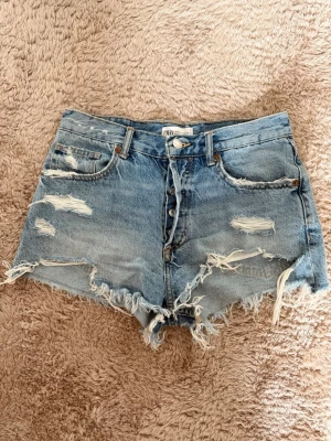 Blå slitna denimshorts från Zara - Säljer ett par blå denimshorts från Zara med hög midja och slitna detaljer. Shortsen har råa kanter, flera rivna partier och klassiska fem fickor. Är lite mer upprivna på ett ställe av misstag men fungerar fortfarande att använda 