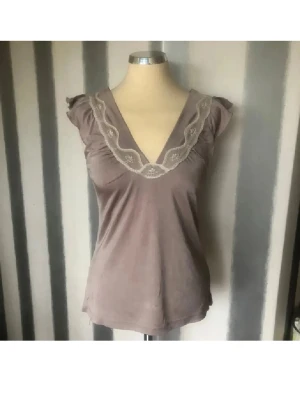 Beige topp med spetsdetaljer - Supersöt beige topp med v-ringning och spetsdetaljer runt halsen. Toppen har korta ärmar med volang och är tillverkad i ett mjukt material som känns skönt mot huden. Perfekt för dig som gillar romantiska och feminina plagg.