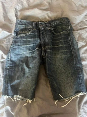 Levi's jeansshorts  - Mörkblå jeansshorts från Levi's och rå, fransig kant nedtill. Shortsen har tydliga slitningar och kontrastsömmar, samt Levi's ikoniska läderpatch bak i midjan. Perfekta för en avslappnad streetstil.