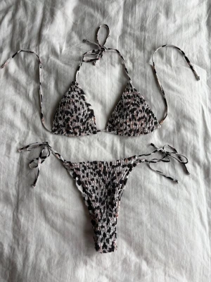 Svart och vit mönstrad bikini - Trendig trekantsbikini med svart, vit och ljusrosa mönster. Både överdel och underdel har smockad struktur och knytband i sidor och nacke för justerbar passform. 