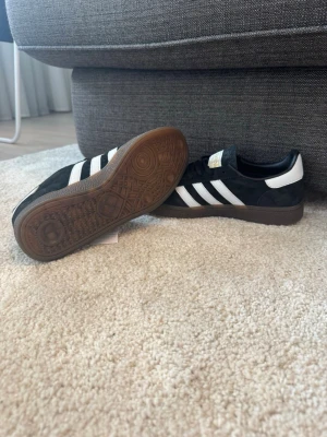Adidas Samba OG svarta sneakers - Adidas Samba OG sneakers i svart mocka med klassiska vita ränder och gummisula. Låg modell med gulddetaljer på plösen och sidan. Perfekta för en stilren och sportig look. Ikonisk design som funkar till det mesta.