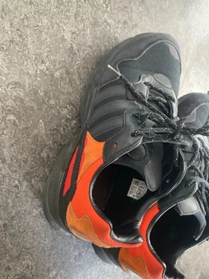 Adidas svarta sneakers med orange detaljer - Säljer ett par svarta Adidas sneakers med snygga orange och röda detaljer på sidorna. Skorna har klassiska tre ränder, svart snörning och en chunky sula. Materialmix av mocka och textil ger en cool look. Perfekta för dig som vill sticka ut lite extra.