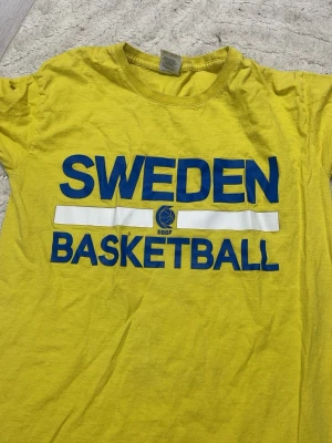Gul Sweden Basketball t-shirt - Gul t-shirt med blå text 'Sweden Basketball' och SBBF-logga på bröstet. Klassisk rund hals och korta ärmar. Perfekt för basketfans som vill visa sitt stöd för Sverige.