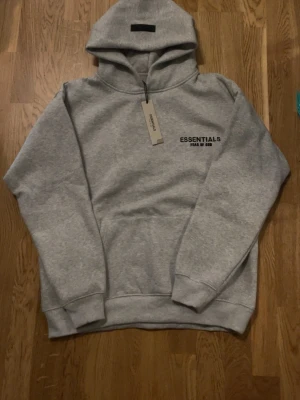 Essentials Fear of God hoodie grå - Pris kan diskuteras.                                             Snygg grå hoodie från Fear of God Essentials med svart logga på bröstet och stor text på ryggen. Klassisk passform med huva och känguruficka. Perfekt för en clean och avslappnad streetwear-look.