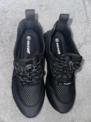Svarta sneakers från Veltuff - Svarta sneakers från Veltuff med sportig design och meshdetaljer för extra andningsförmåga. Skorna har svarta och vita snören, dragflik baktill och en låg siluett. Perfekta för en stilren och bekväm look.