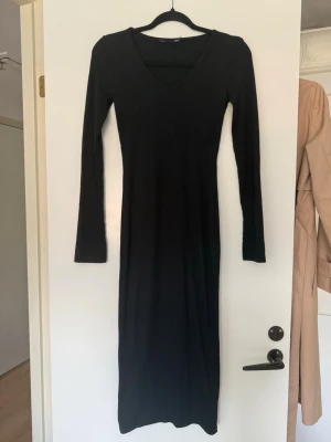 Svart ribbad långklänning - Svart långklänning med v-ringning och lång ärm. Klänningen är ribbad och har en smickrande, kroppsnära passform. Perfekt för dig som gillar en stilren och enkel look.