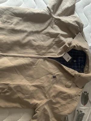 Beige jacka från Polo Ralph Lauren - Snygg beige jacka från Polo Ralph Lauren med dragkedja och klassisk krage. Jackan har ett rutigt innerfoder i marinblått och vitt samt broderad logga på bröstet. Perfekt för dig som vill ha en stilren och tidlös look.