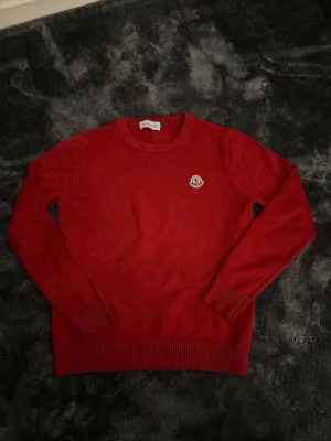 Röd Moncler tröja med logga - Säljer en röd tröja från Moncler med rund halsringning och ribbade muddar. Tröjan har en broderad Moncler-logga på bröstet och är långärmad. Perfekt för dig som gillar klassisk och stilren design.