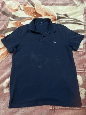 Marinblå pikétröja från GANT - Klassisk marinblå pikétröja från GANT med korta ärmar och krage. Tröjan har knappar vid halsen och en liten broderad GANT-logga på bröstet. Perfekt för en stilren och avslappnad look.