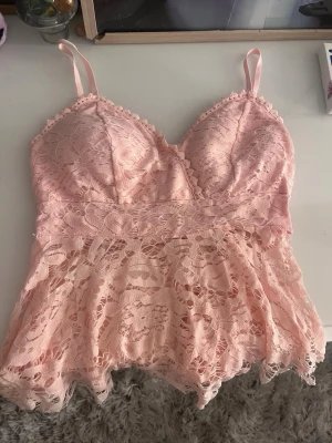 Rosa spetsbody med tunna axelband - Söt rosa body i spets med tunna axelband och v-ringning. Modellen har vadderade kupor och ett fint blommigt spetsmönster över hela plagget. Nederdelen är lätt utsvängd och ger en romantisk känsla. Perfekt för dig som gillar feminina detaljer.