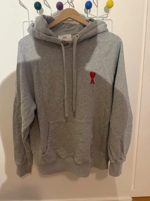 Grå hoodie från Ami Paris - Snygg grå hoodie från Ami Paris med klassisk huva och dragsnören. På bröstet sitter Ami:s ikoniska röda hjärta med A-broderi. Hoodien har en stor magficka och ribbade muddar vid ärmar och nederkant. Perfekt för en chill och stilren look.