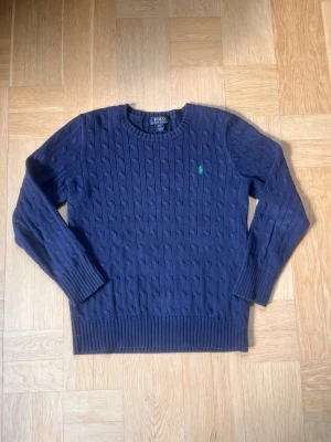 Mörkblå kabelstickad tröja Polo - Mörkblå kabelstickad tröja från Polo Ralph Lauren med rund halsringning och grönt broderat logomärke på bröstet. Tröjan har långa ärmar och ribbade muddar vid ärmslut och nederkant. Perfekt för en stilren och klassisk look.