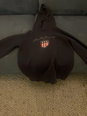 Svart hoodie från GANT med logga - Svart hoodie från GANT med klassisk huva och stor broderad logga samt sköld på bröstet. Hoodien har långa ärmar och en avslappnad passform, perfekt för en chill look. Materialet är mjukt och bekvämt, troligen bomull eller bomullsblandning.