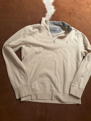 Beige tröja med half zip från Polo Ralph Lauren - Snygg beige långärmad tröja från Polo Ralph Lauren med half zip och klassisk logga på bröstet. Tröjan har en avslappnad passform och är tillverkad i mjuk bomull, perfekt för en stilren vardagslook.