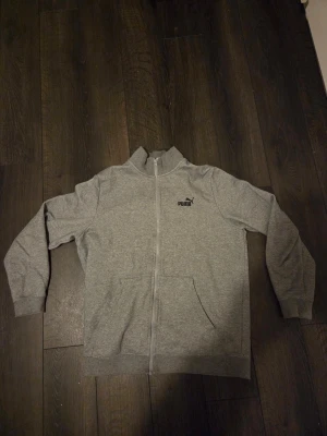 Grå zip-hoodie från Puma - Snygg grå zip-hoodie från Puma med svart logga på bröstet. Tröjan har dragkedja hela vägen, två fickor fram och ribbade muddar vid ärmslut och midja. Perfekt för en avslappnad och sportig stil.