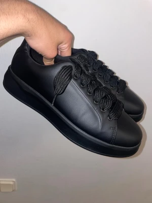 ZARA Svarta chunky sneakers i läderlook  - Stilrena svarta sneakers med chunky sula och breda svarta skosnören. Skorna har en clean design i läderimitation och passar perfekt till streetwear eller casual outfits. Lågt skaft och helsvart look ger en modern vibe. Skor från Zara. Storlek 43. Får ett extra par sko snören om du vill byta ut dem tjocka. 