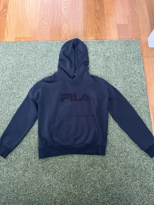 Svart hoodie från FILA - Svart hoodie från FILA med stor logga framtill. Klassisk modell med huva och magficka. Mjuk bomullsblandning och ribbade muddar vid ärmslut och nederkant. Perfekt för en avslappnad och sportig stil. Storleken e inte riktigt passformen till L passar mer M kanske S.