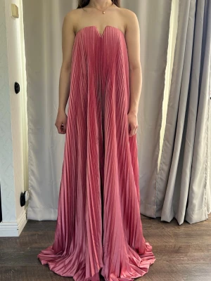 Axelbandslös plisserad maxiklänning - Säljer en axelbandslös maxiklänning i plisserat tyg med vacker rosa ton. Klänningen har hjärtformad urringning och ett elegant, flytande fall. Perfekt för dig som vill sticka ut med en unik och trendig look. Klänningen är i helt nytt skick och lapparna sitter fortfarande kvar. 