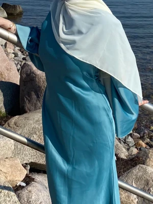 Lång blå klänning med vida ärmar - Elegant långklänning i klarblått med vida, fladdrande ärmar och enkel, slät design. Klänningen har en figurnära passform och är tillverkad i ett glansigt material som ger ett exklusivt intryck. Perfekt för dig som vill sticka ut med stilren look. Passar XS, Soch M