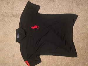 Svart pikétröja från Polo Ralph Lauren - m - polyester - Svart pikétröja från Polo Ralph Lauren med stor röd logga på bröstet och röd siffra på ärmen. Klassisk krage och knäppning framtill. Perfekt för en stilren och sportig look. I polyester