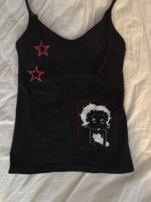 Svart linnetopp med stjärnor och tryck - Svart linnetopp med tunna axelband, två röda stjärnor på bröstet och ett vitt tryck av betty boop. Toppen har en enkel, tight passform och är perfekt för en cool och personlig stil. Jag har målat den själv, och det är ett vanligt linne från lager 157 egentligen. Storlek S