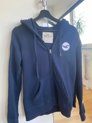 Mörkblå hoodie från Hollister - Mörkblå zip-hoodie från Hollister med vit logga på bröstet och tryck på huvan. Hoodien har dragsko i huvan, två fickor fram och ribbade muddar. Perfekt för en avslappnad och cool stil.