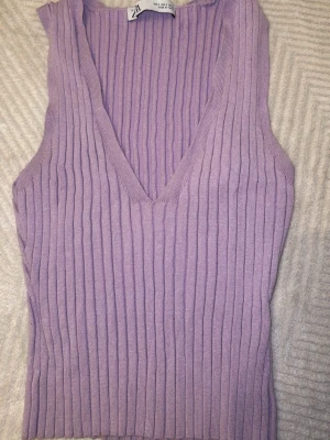 Lila ribbad stickad linnetopp Zara - Superfin lila stickad linnetopp från Zara med v-ringning och ribbad struktur. Perfekt att styla med jeans eller kjol för en trendig look. Ärmlös och mjuk i materialet.