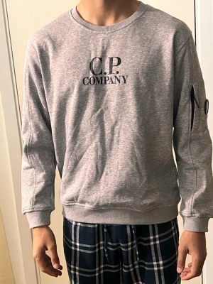 Grå sweatshirt från C.P. Company - Grå sweatshirt från C.P. Company med svart logga på bröstet. Tröjan har rund halsringning, långa ärmar och en ficka med dragkedja på ena ärmen. Tillverkad i mjuk bomull och har ribbade muddar vid ärmslut och nederkant.