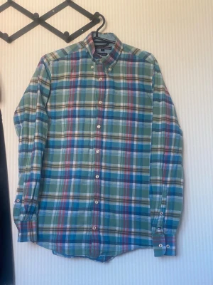 Rutig skjorta i bomull från Tommy Hilfiger - Säljer en klassisk rutig skjorta från Tommy Hilfiger i mjuk bomull. Skjortan har blå, gröna, röda och vita rutor, knappar framtill och på kragen samt långa ärmar med knapp vid manschetten. Perfekt för en avslappnad men snygg stil.