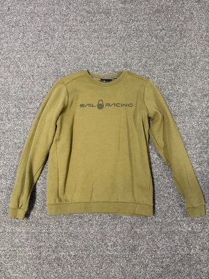 Olivgrön sweatshirt från Sail Racing - Snygg olivgrön sweatshirt från Sail Racing med tryckt logga på bröstet och liten logga i nacken. Tröjan har rund hals, långa ärmar och ribbade muddar vid ärmslut och nederkant. Perfekt för en avslappnad och sportig stil. Storlek 170 använd 1-2 gånger