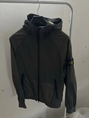 Mörkgrön vindjacka från Stone Island - Snygg mörkgrön vindjacka från Stone Island med ikonisk patch på vänster ärm. Jackan har huva, dragkedja framtill och två fickor med dragkedja. Tillverkad i lätt, vindtätt material som passar perfekt för blåsiga dagar.