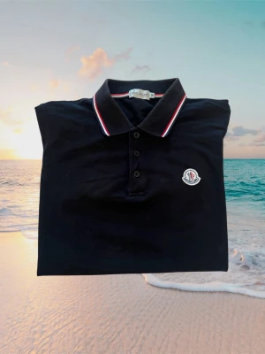 Svart Moncler pikétröja med rödvit krage - Klassisk svart pikétröja från Moncler med rödvita detaljer på krage och ärmslut. Tröjan har knappslå framtill och Moncler-logga på bröstet. Tillverkad i mjuk bomull och har en normal passform.