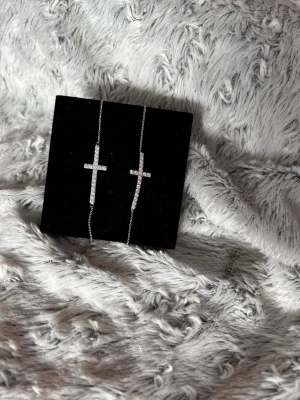 Edblad: Silverarmband och silverhalsband med kors och stenar - tunna silverfärgade armband och halsband med korsberlocker täckta av små gnistrande stenar. Smyckena har en minimalistisk och stilren design som passar perfekt till en trendig look. Kedjan är tunn och smidig, vilket gör dem lätta att bära tillsammans eller separat.