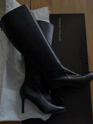Svarta läderstövlar från Bottega Veneta - Snygga svarta knähöga stövlar från Bottega Veneta i slätt läder. Stövlarna har spetsig tå och unik flätad klack som ger en cool touch. Dragkedja baktill för enkel påtagning. Perfekta för dig som vill ha en stilren och trendig look.
